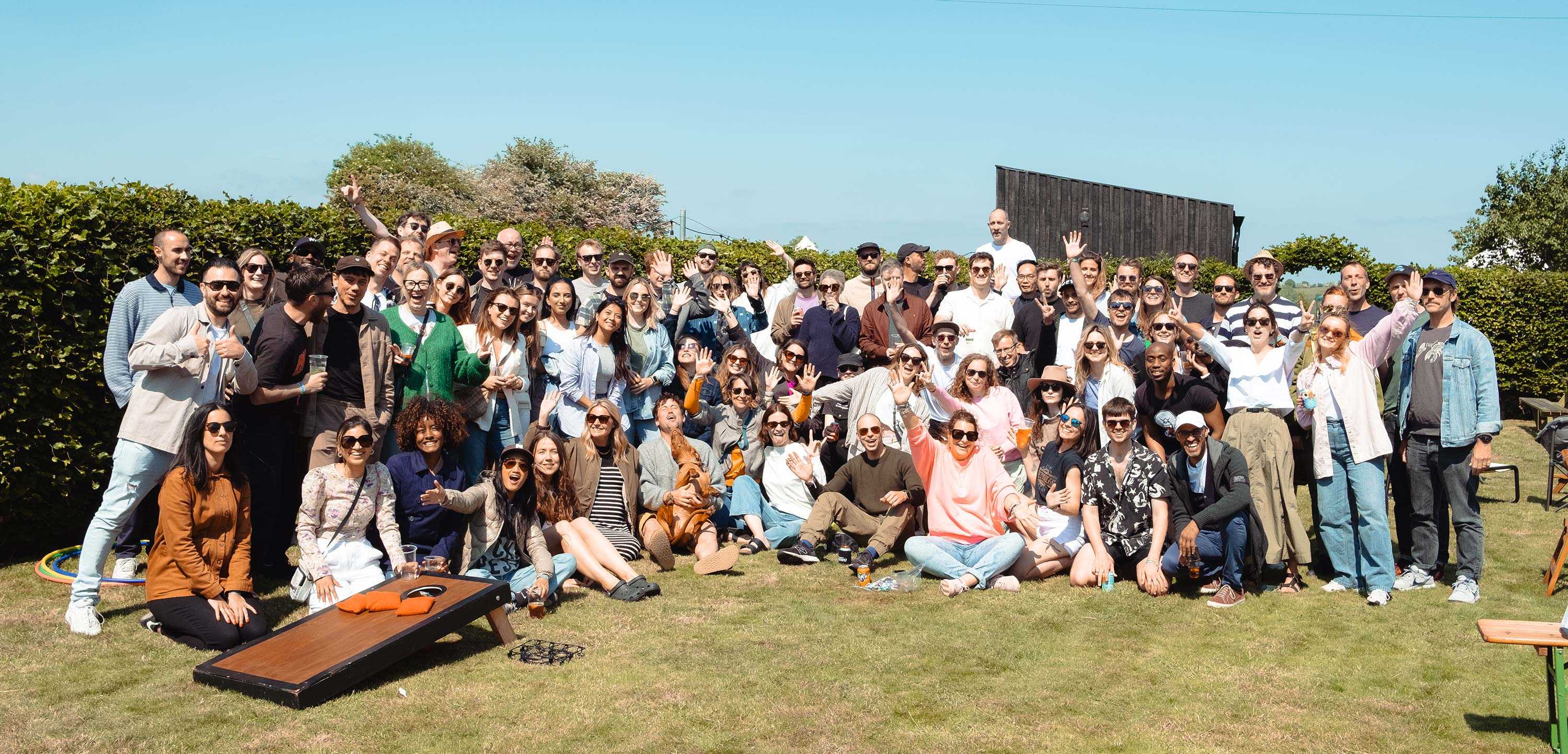 A group photo of employees at our annual social day - smiling, waving and cheering against a bucolic scene of green grass and blue sky